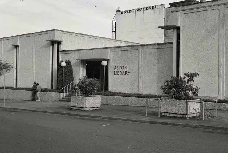 A black and white photograph of the street entrance to the Astoria Public Library - at the time known as the Astor Library. The sign near the entrance reads, "Astor Library". Behind the library there is a building that reads, "Hotel Waldorf".